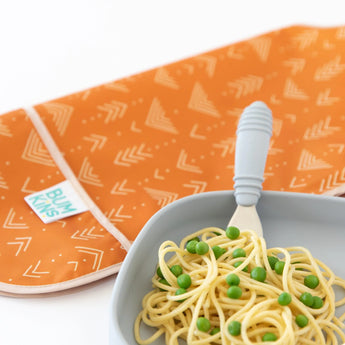 Close-up of a gray plate of spaghetti and peas and a gray fork, with a folded Bumkins Junior Bib: Grounded behind on a white background.