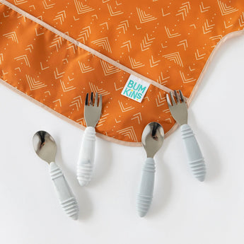 Close-up of two sets of children's utensils, with Bumkins Junior Bib: Grounded partially visible on a white background.