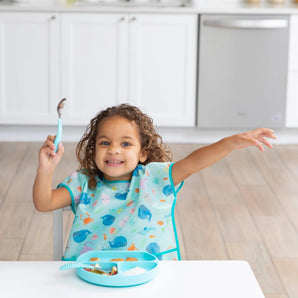 A curly-haired child in a Bumkins Junior Bib: Ocean Life sits at a table with a blue plate, holding a spoon and raising an arm excitedly.