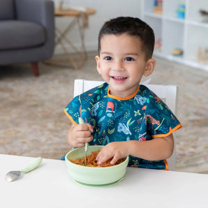A child in a Bumkins Junior Bib: Jungle smiles, enjoying pasta from a green bowl at a white table.