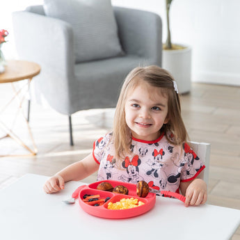 Young girl beams at table in a Minnie Mouse shirt with her red plate, wearing a Bumkins Junior Bib: Minnie Mouse Classic for easy cleanup.