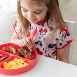A girl in a Minnie Mouse shirt enjoys eggs, fruit, and meatballs from a red plate, wearing her Bumkins Junior Bib: Minnie Mouse Classic.