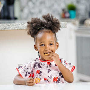 A curly-haired child in Bumkins Junior Bib: Minnie Mouse Classic smiles, eating a pretzel at a white table in a kitchen.