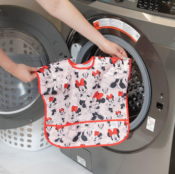 Someone is putting a Bumkins Junior Bib: Minnie Mouse Classic, with an easy-wipe stain-resistant design, into the washing machine.