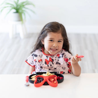 A girl with long hair enjoys fruit at a table, wearing a Bumkins Junior Bib: Minnie Mouse Classic. A potted plant complements the scene.