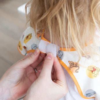 Close-up of hands fastening the hook-and-loop closure of a Bumkins Junior Bib: Winnie and Friends around a child's neck. 