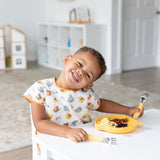A child wearing a Bumkins Junior Bib: Winnie and Friends eats from a yellow plate at a white table in a bright room.