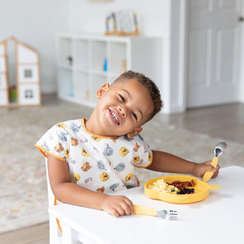 A child wearing a Bumkins Junior Bib: Winnie and Friends eats from a yellow plate at a white table in a bright room.