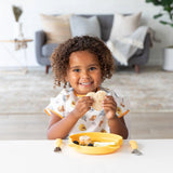 A child beams, holding a sandwich by a yellow plate and utensils, wearing a Bumkins Junior Bib: Winnie and Friends in a cozy living room.