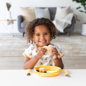 A child beams, holding a sandwich by a yellow plate and utensils, wearing a Bumkins Junior Bib: Winnie and Friends in a cozy living room.