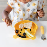 A child eats sandwiches, berries, and celery from a bear-shaped plate while wearing a Bumkins Junior Bib: Winnie and Friends at a table.