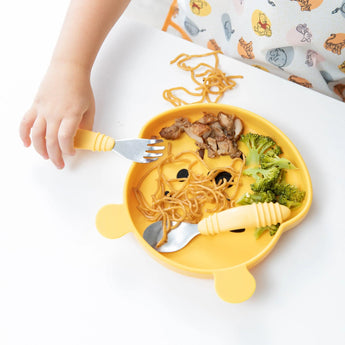 Close-up of a child wearing Bumkins Junior Bib: Winnie and Friends, holding a fork near a bear-shaped plate of food on a white table.