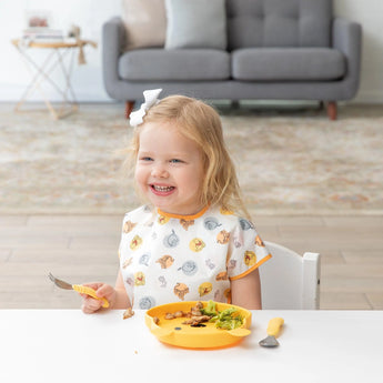 A girl in a Bumkins Junior Bib: Winnie and Friends smiles while eating with a fork from a yellow plate at a white table.