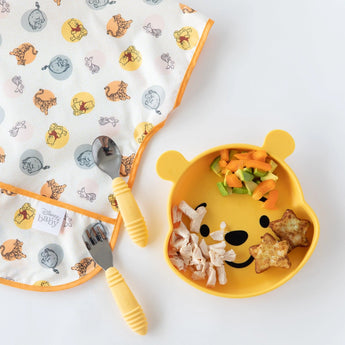 Bumkins Junior Bib: Winnie and Friends lays flat next to a bear-shaped plate of food and yellow utensils on a white background.