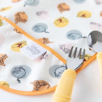 Close-up of a yellow fork and spoon lying on top of Bumkins Junior Bib: Winnie and Friends on a white background.