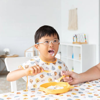 A child with glasses enjoys a meal from a yellow plate with an adult nearby, wearing a Bumkins Junior Bib: Winnie and Friends.