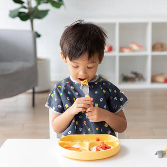 Child in a Bumkins Junior Bib: Super Mario™ Lineup eats from a yellow plate using a fork at a table in a bright room with shelves.