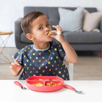 A young child enjoys chicken nuggets on a red plate at a white table, wearing a Bumkins Super Mario™ Lineup Junior Bib by a cozy couch.