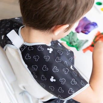 A child painting while wearing a Bumkins Short-Sleeved Smock: Mickey Mouse Icon B+W, with the hook-and-loop neck closure visible.