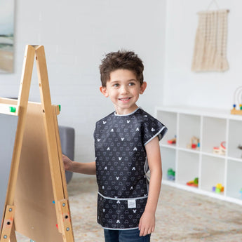 A boy in a Bumkins Short-Sleeved Smock: Mickey Mouse Icon B+W stands by an easel, smiling and ready to paint in a playroom.