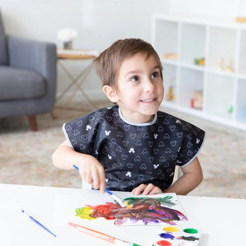 A child painting with watercolors at a white table, wearing a Bumkins Short-Sleeved Smock: Mickey Mouse Icon B+W in a cozy room.