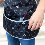 Close-up of a child in a Bumkins Short-Sleeved Smock: Mickey Mouse Icon B+W, with paintbrushes in the pocket.