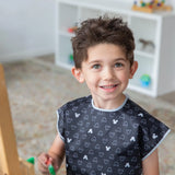 A child smiles, painting in a Bumkins Short-Sleeved Smock: Mickey Mouse Icon B+W; toys and books fill the shelves behind.