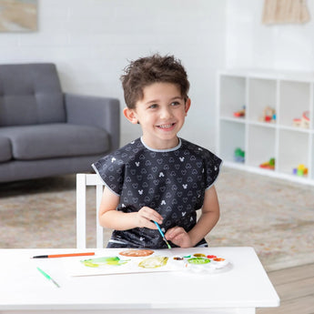A child paints at a white table in a bright room, wearing a Bumkins Short-Sleeved Smock: Mickey Mouse Icon B+W, near a gray sofa and shelf.