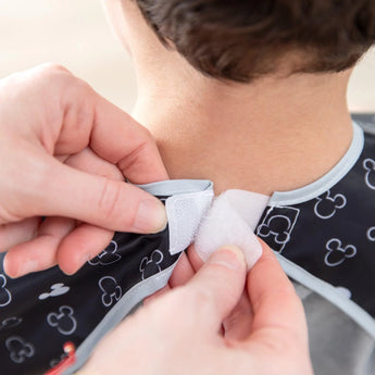 Close-up of hands securing the hook-and-loop closure of a Bumkins Short-Sleeved Smock: Mickey Mouse Icon B+W around a child's neck.
