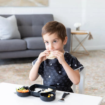 A child wearing a Bumkins Short-Sleeved Smock: Mickey Mouse Icon B+W enjoys a sandwich with food on a sectioned tray at a white table.