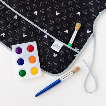 Close-up of a Bumkins Short-Sleeved Smock: Mickey Mouse Icon B+W on a white background, with a paint palette and paintbrushes nearby.