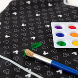 Close-up of a paint brush and paint palette on top of a Bumkins Short-Sleeved Smock: Mickey Mouse Icon B+W, with a smear of green paint.