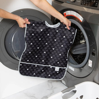 A person holds a Bumkins Short-Sleeved Smock: Mickey Mouse Icon B+W in front of an open front-loading washing machine.