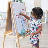A child in a Bumkins Short-Sleeved Smock: Mickey Mouse Classic paints vibrant strokes at an easel on wooden floors in a bright room.