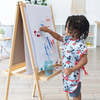 A child in a Bumkins Short-Sleeved Smock: Mickey Mouse Classic paints vibrant strokes at an easel on wooden floors in a bright room.