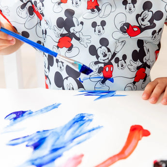 Close-up of a child painting in blue, wearing a Bumkins Short-Sleeved Smock: Mickey Mouse Classic.