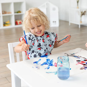 A child with curly blonde hair paints at a white table in a bright room, wearing a Bumkins Short-Sleeved Smock: Mickey Mouse Classic.