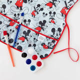 Close-up of Bumkins Short-Sleeved Smock: Mickey Mouse Classic on a white background, next to a paint palette and paintbrushes.