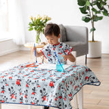 A child paints at a table in a Bumkins Short-Sleeved Smock: Mickey Mouse Classic, amidst a blue water jar, art supplies, and a potted plant.