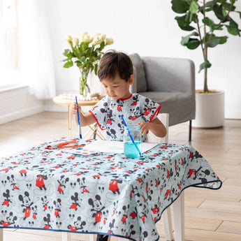 A child paints at a table in a Bumkins Short-Sleeved Smock: Mickey Mouse Classic, amidst a blue water jar, art supplies, and a potted plant.