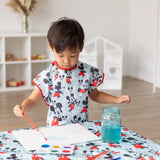 A child paints at a table wearing a Bumkins Short-Sleeved Smock: Mickey Mouse Classic, with a palette and a jar of water nearby.