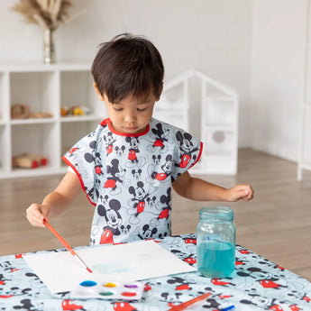 A child paints at a table wearing a Bumkins Short-Sleeved Smock: Mickey Mouse Classic, with a palette and a jar of water nearby.