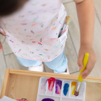 A child paints joyfully with colorful paints, protected by a Bumkins Short-Sleeved Smock: Princess Magic for easy cleaning.