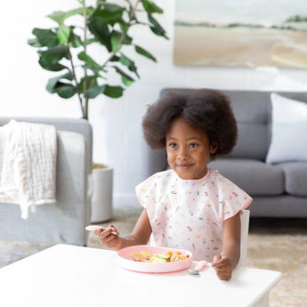 A child with curly hair enjoys a meal from a pink plate, wearing a Bumkins Short-Sleeved Smock: Princess Magic in a bright living room.