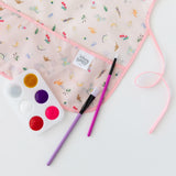 Close-up of Bumkins Short-Sleeved Smock: Princess Magic on a white background next to paintbrushes and a paint palette.