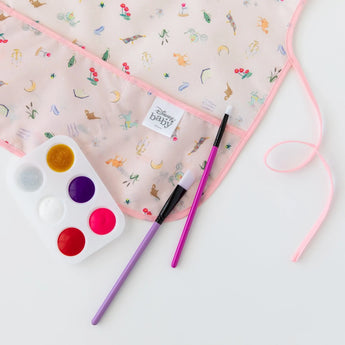 Close-up of Bumkins Short-Sleeved Smock: Princess Magic on a white background next to paintbrushes and a paint palette.