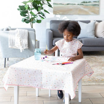 In a cozy living room, a child in a Bumkins Short-Sleeved Smock: Princess Magic paints at a table next to a jar of water.