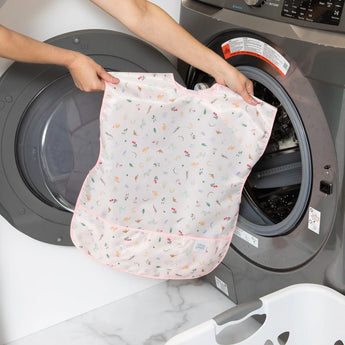 A person holds a Bumkins Short-Sleeved Smock: Princess Magic in front of a front-loading washing machine.
