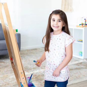 A young girl beams as she paints at an easel, wearing a Bumkins Short-Sleeved Smock: Princess Magic.