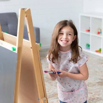 A girl with a paintbrush smiles in a bright room, wearing a Bumkins Short-Sleeved Smock: Princess Magic as she paints at an easel.